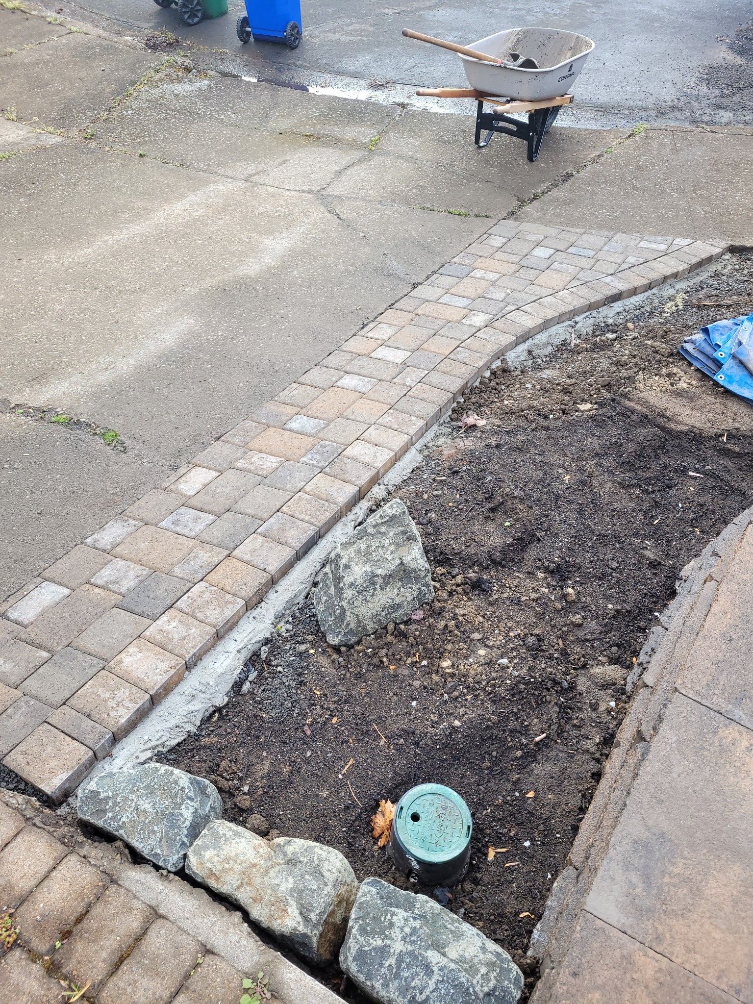 Garden bed beside a wet paved path with soil, stones, and a small green sprinkler head