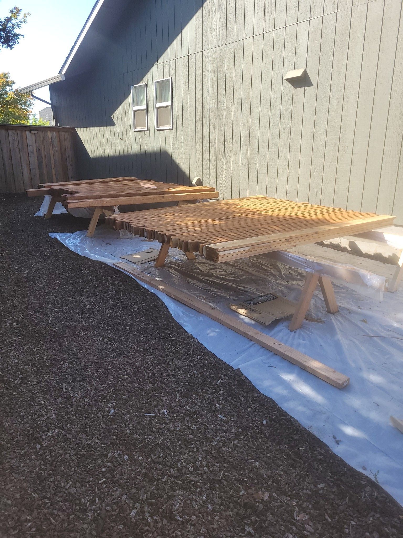 Outdoor work area beside a house with two wooden picnic tables on a tarp over gravel.