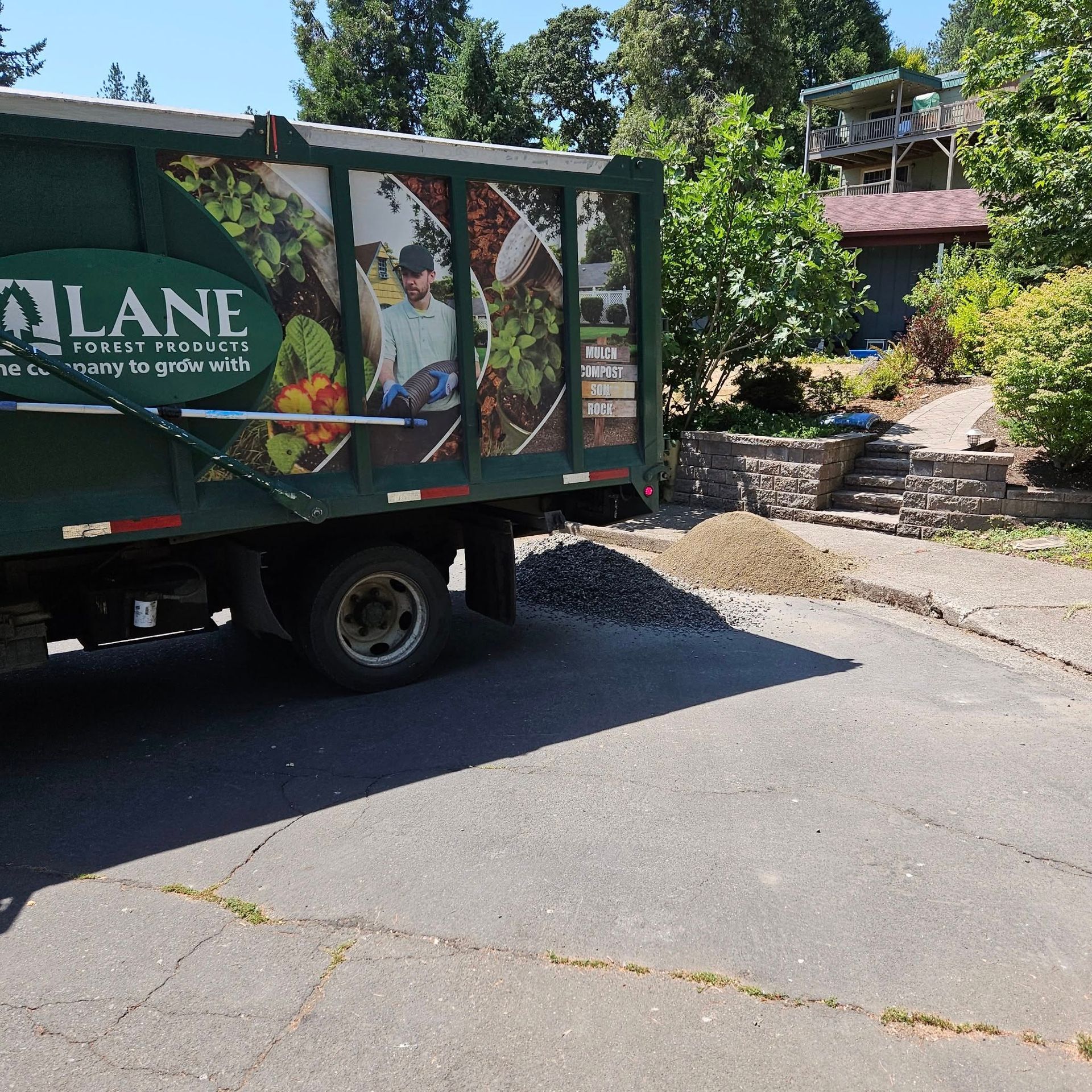 Green landscape truck parked by a driveway beside a house and garden in bright sunlight