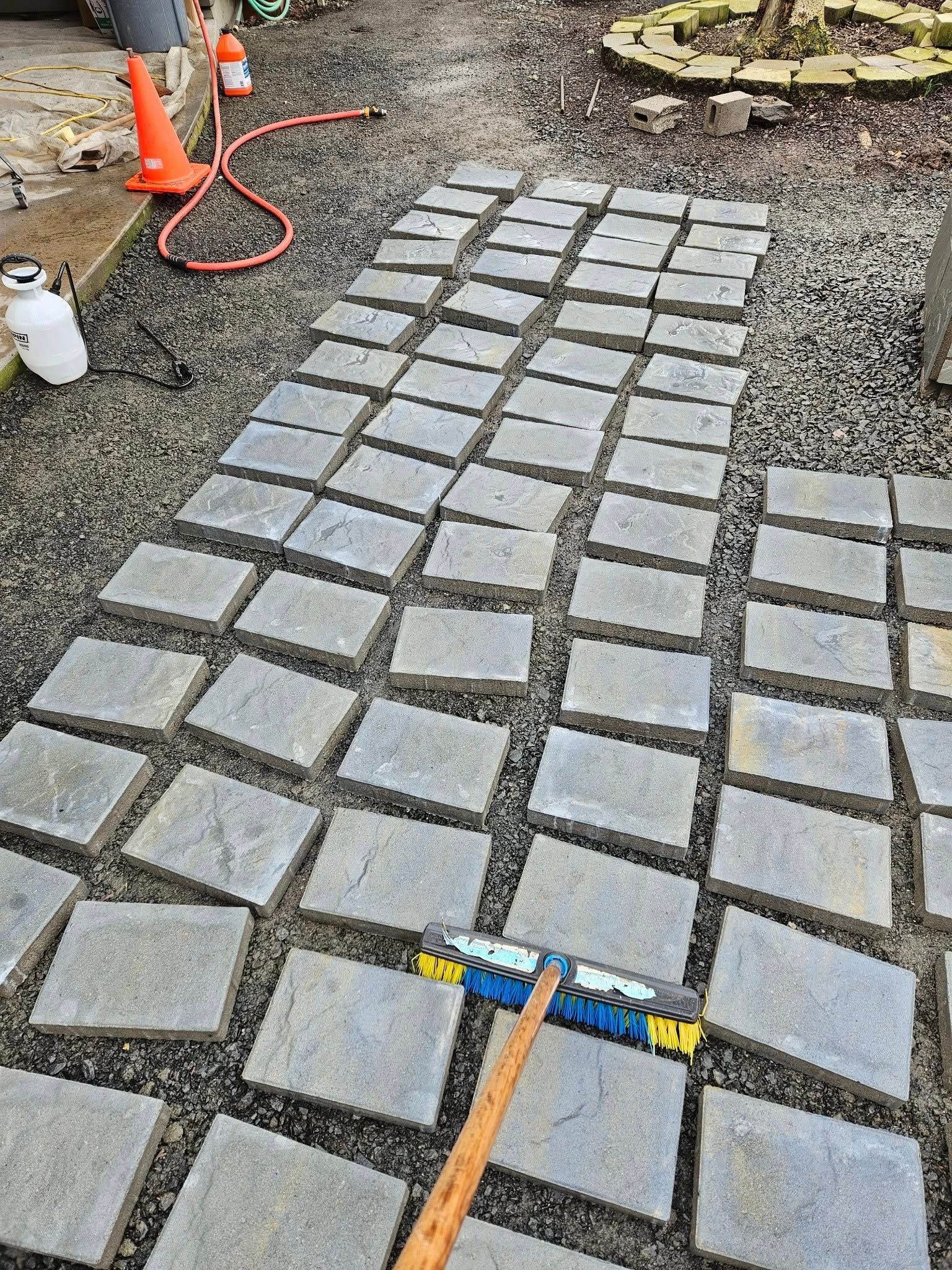 Stone pavers being laid in a gravel path, with tools and orange cones at the edge of a garden area