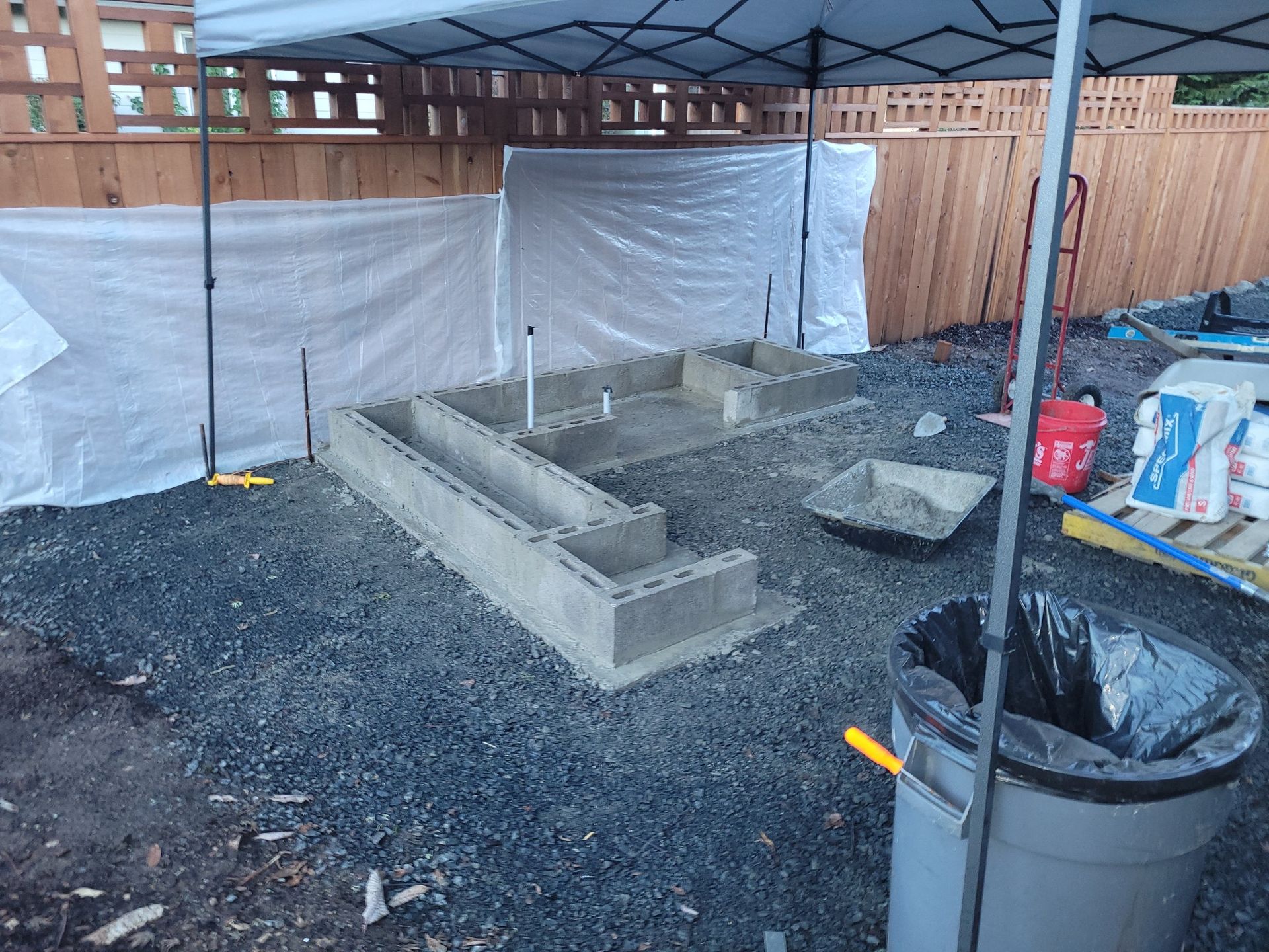 Backyard concrete block planter under a metal canopy, with gravel, fence, and construction materials nearby