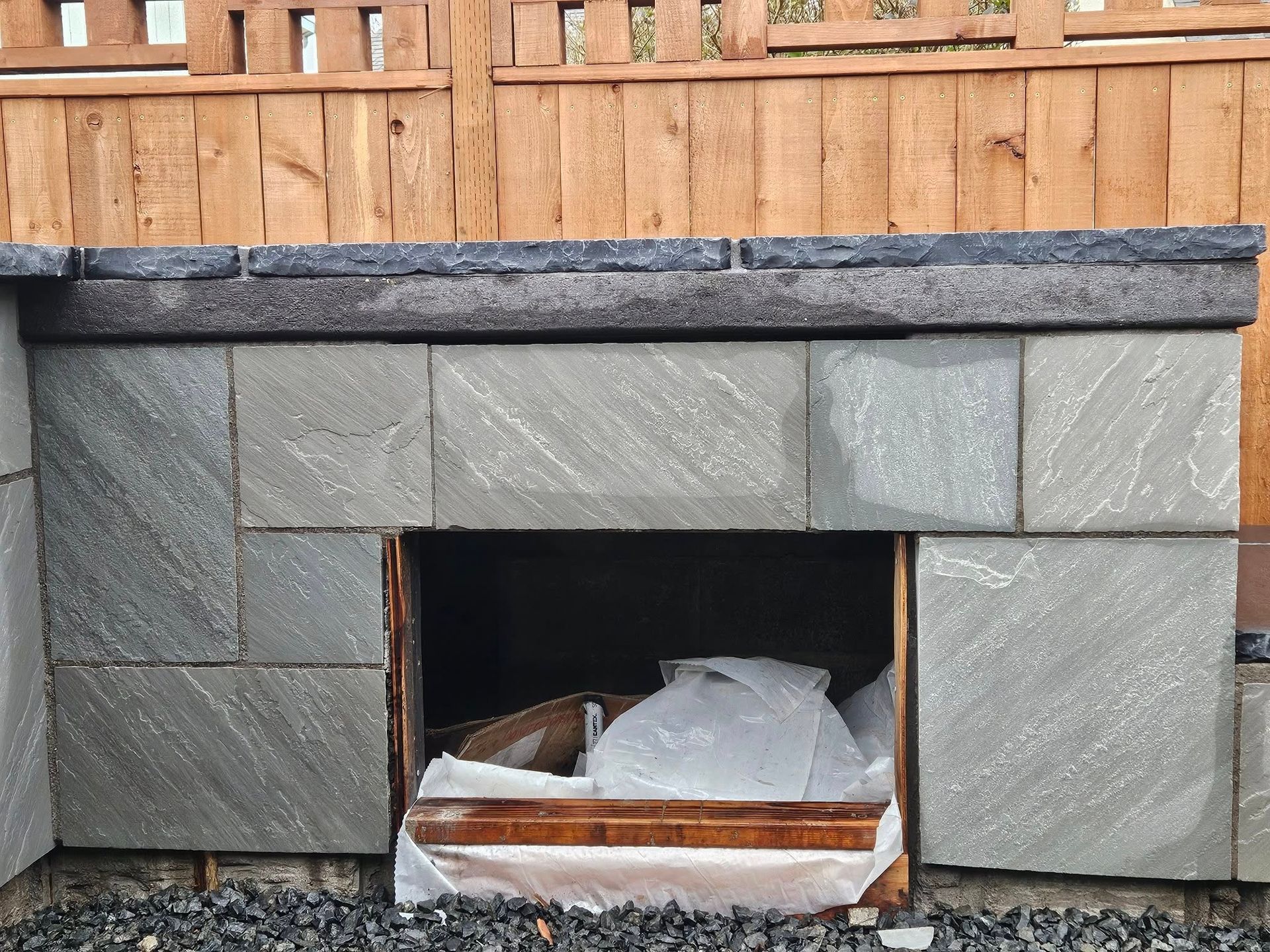 Stone outdoor fireplace with a black mantel and wood fence above it, opening filled with plastic-covered debris