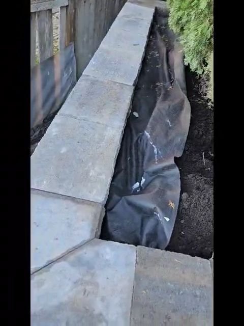 Concrete walkway beside a narrow trench covered with black plastic sheeting and rocks.