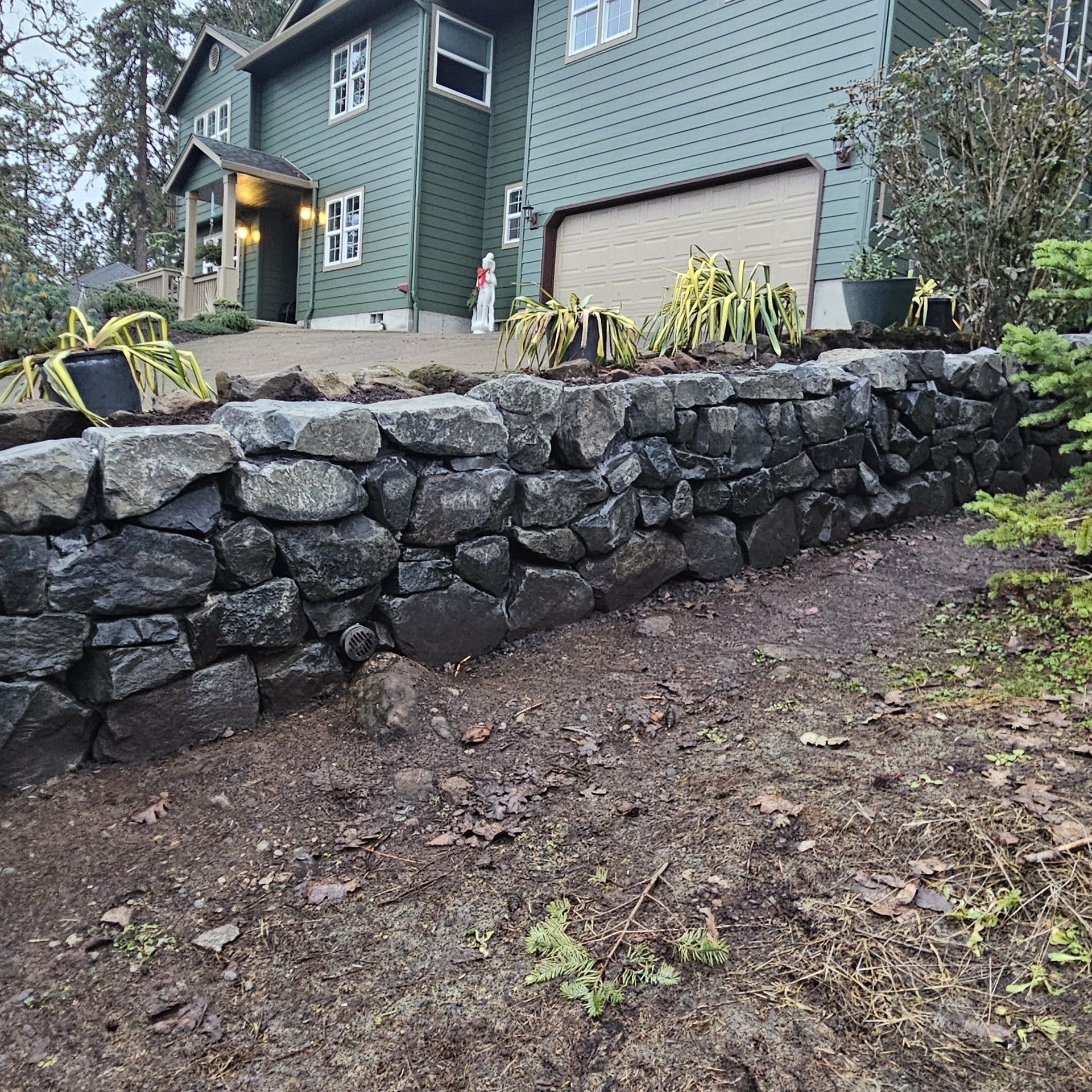 Stone retaining wall beside a green house with a garage, overlooking a dirt slope and landscaped plants