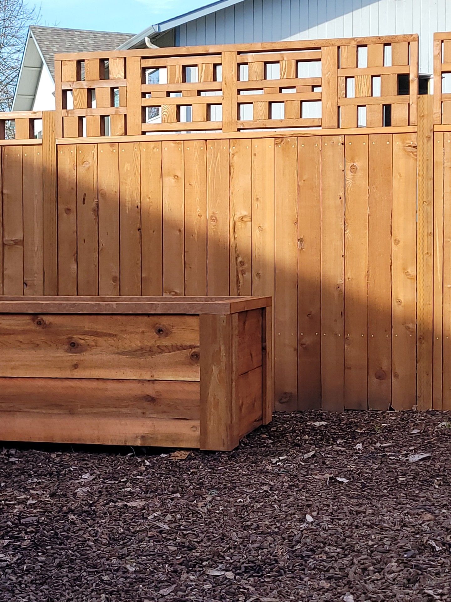 Wooden privacy fence and raised planter in a mulched backyard, with house rooftops visible above.