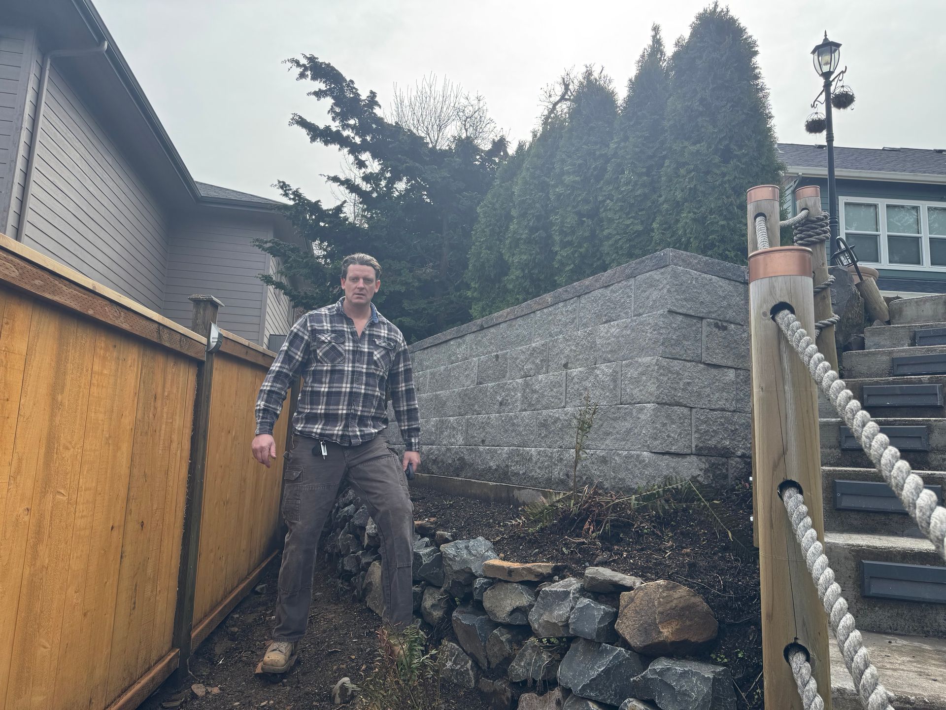 Man standing in a backyard beside a wooden fence and stone retaining wall, with stairs and rope railing nearby