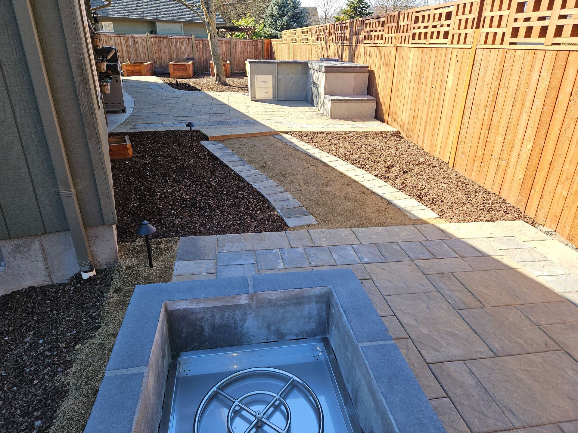 Backyard with a square in-ground fire pit, paver walkway, gravel, and a wooden fence under construction