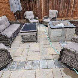 Patio with wicker chairs, cushioned seating, and fire tables on stone pavers, against a wooden fence.