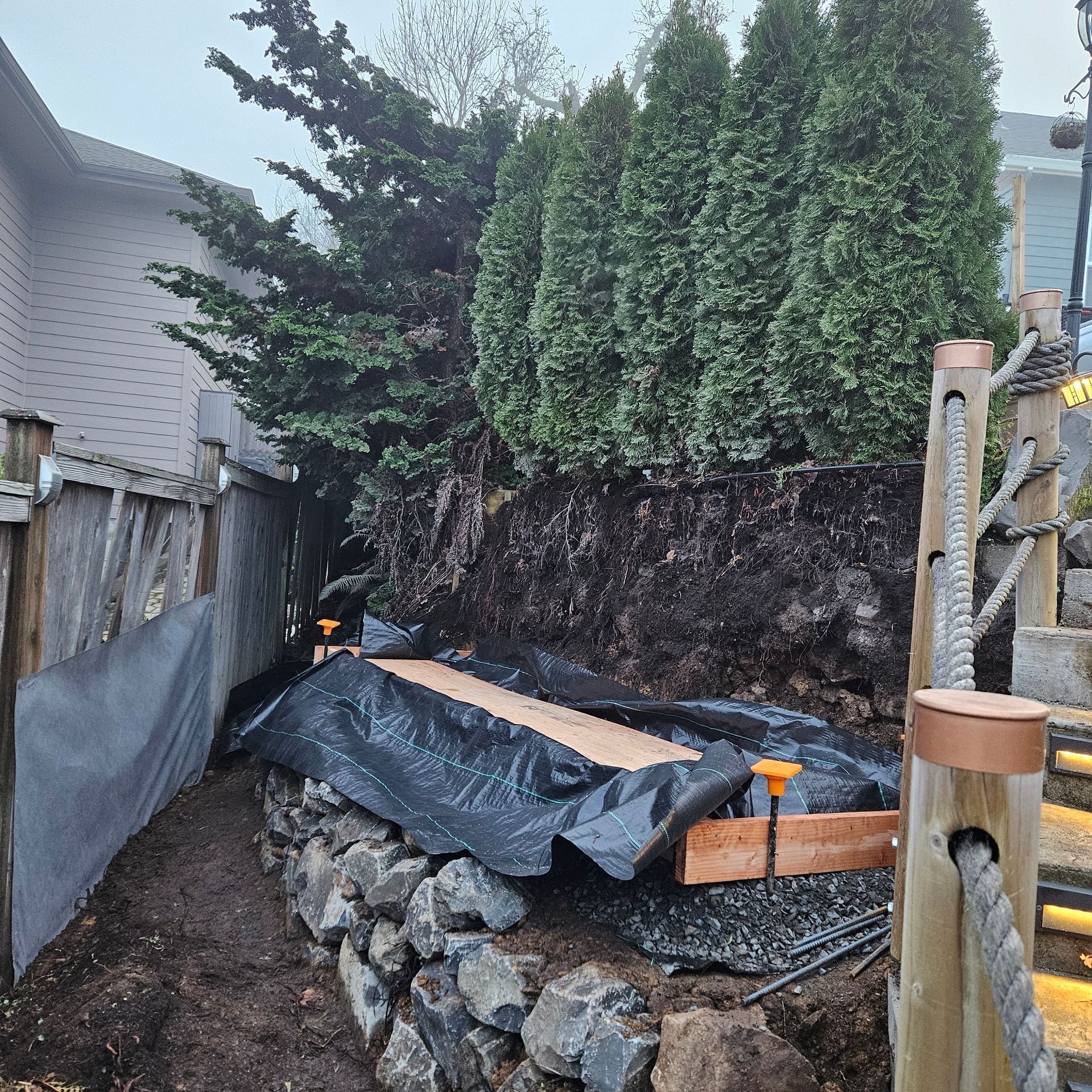 Backyard retaining wall under construction beside a wooden fence and shrubs, with black tarp and stacked rocks