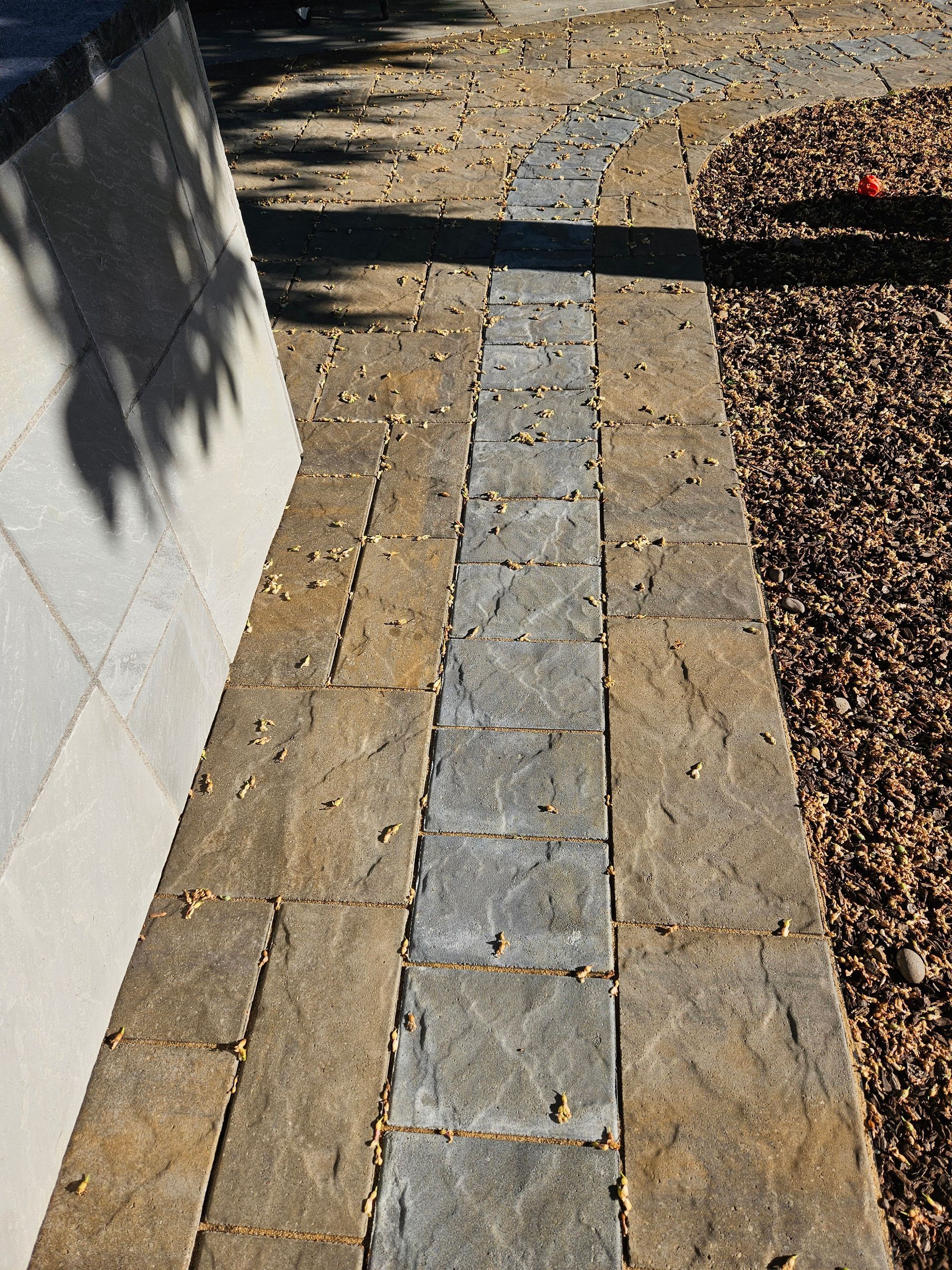 Narrow stone walkway with a gray center strip beside a white wall and brown gravel bed in sunlight