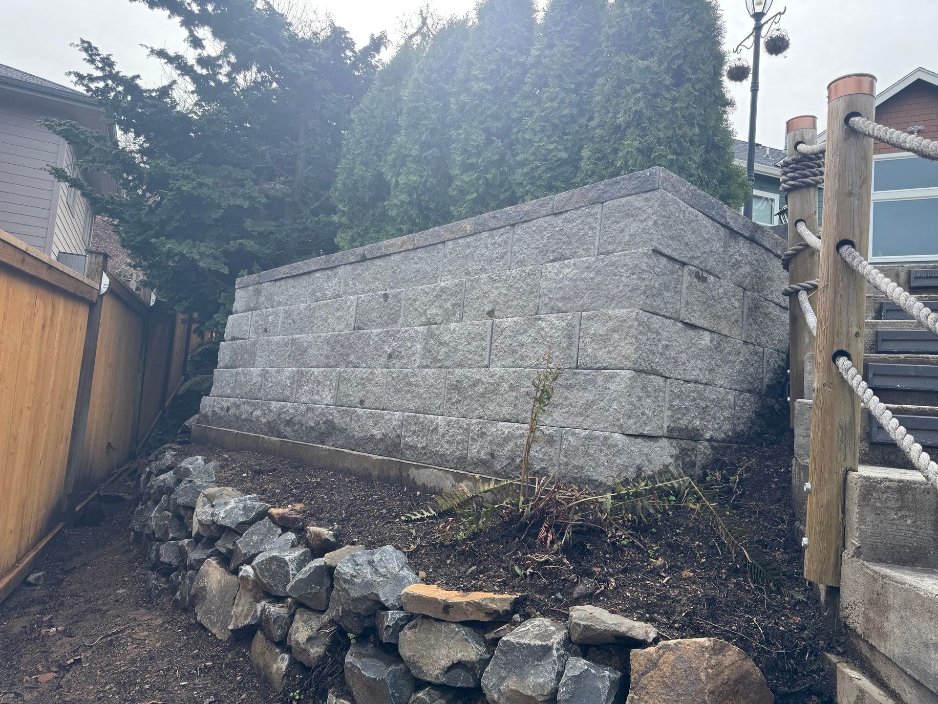 Stone retaining wall in a narrow outdoor path beside a wooden fence and house, with steps on the right.