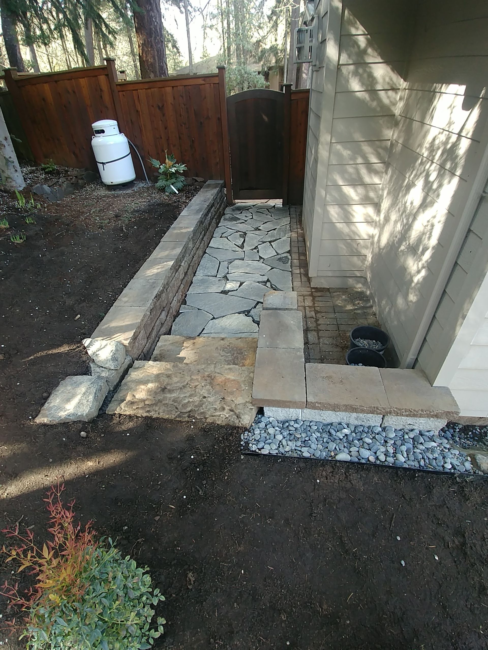 Narrow stone walkway between a house and fence, leading to a dark gate in a shaded backyard.
