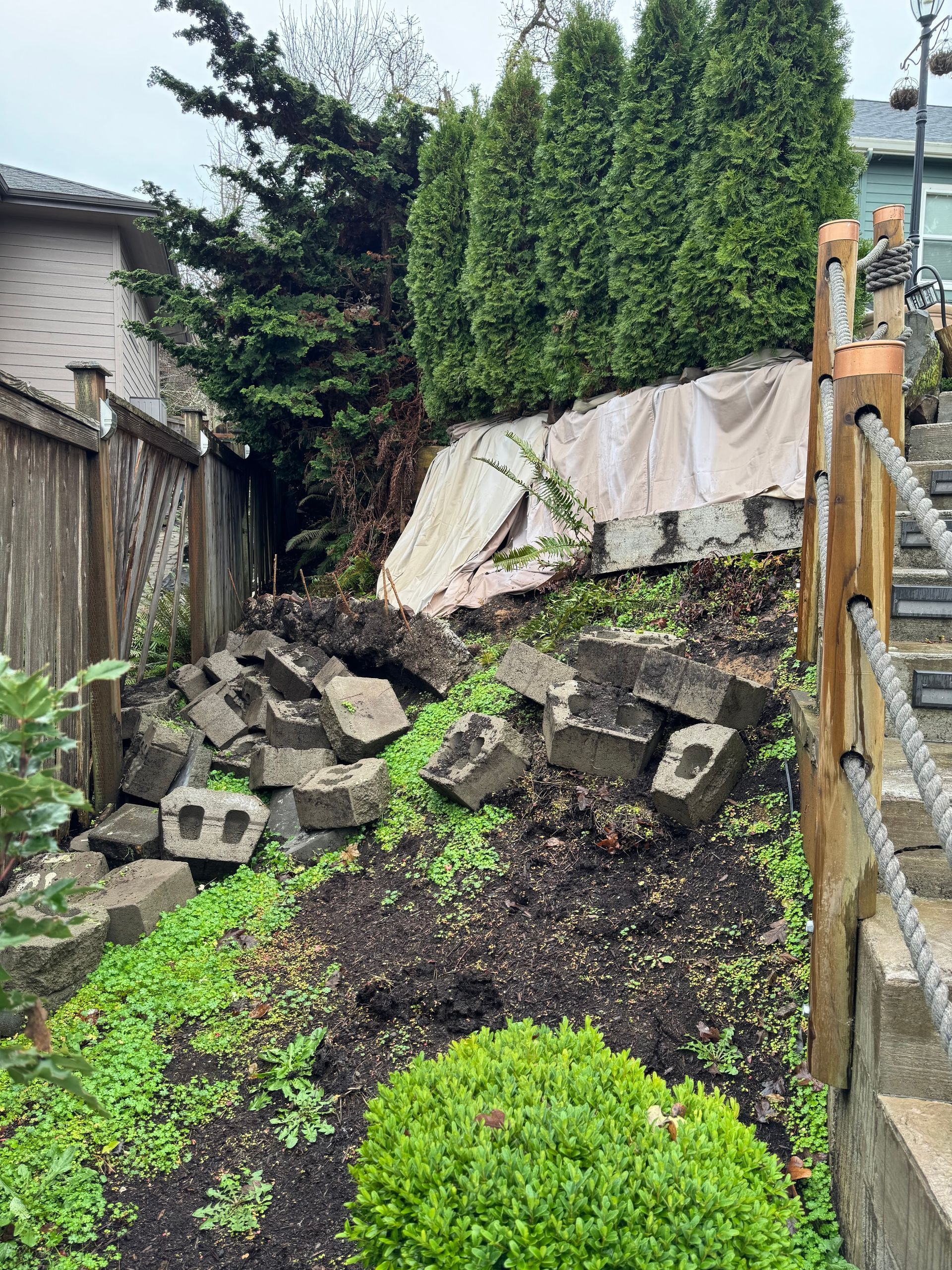Collapsed backyard retaining wall with scattered concrete blocks, wet soil, and greenery after a landslide