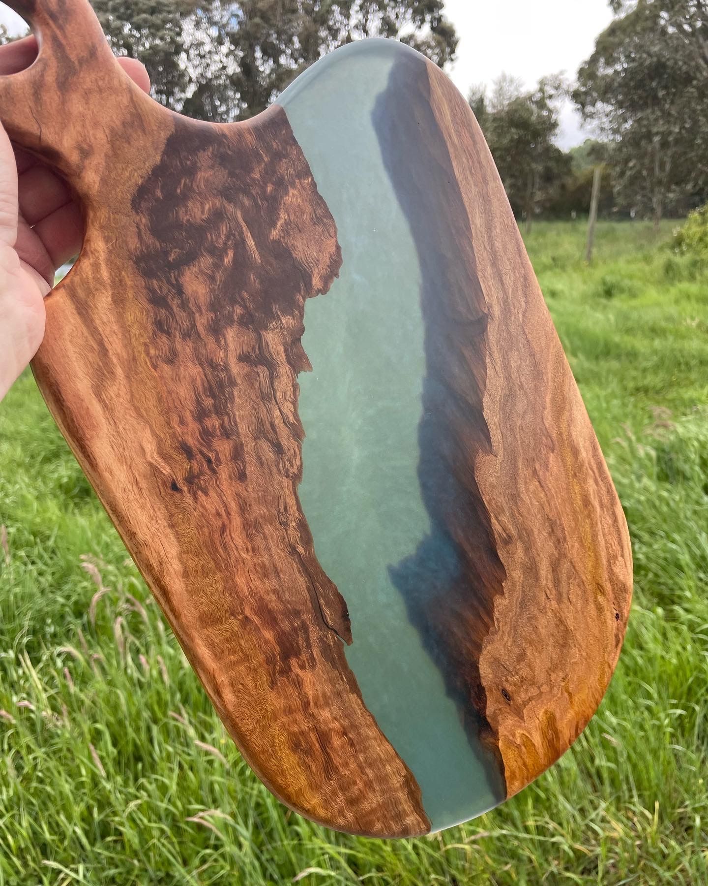 Wooden cutting board with teal resin river detail, held outdoors in grassy field.