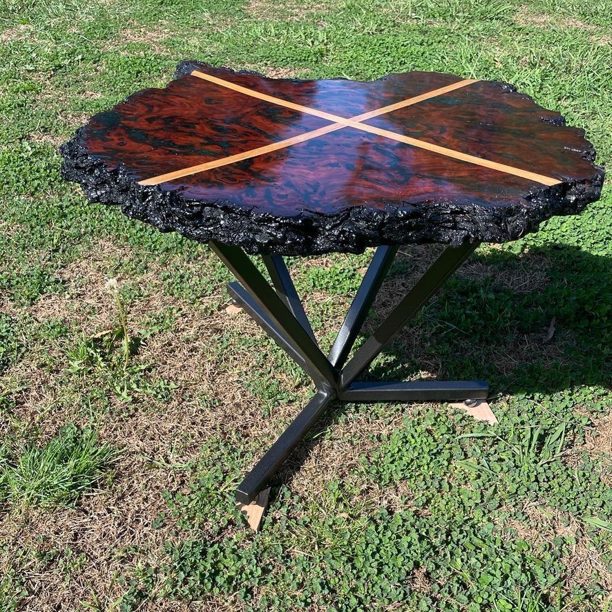 Live-edge wood table with an X-shaped inlay and a black metal base, set on grass.