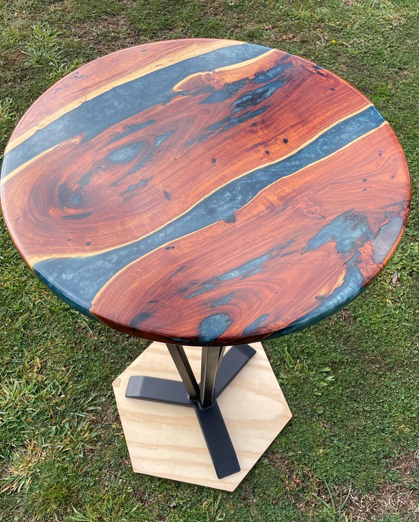Round wooden table with blue resin river, black metal base, and plywood platform.