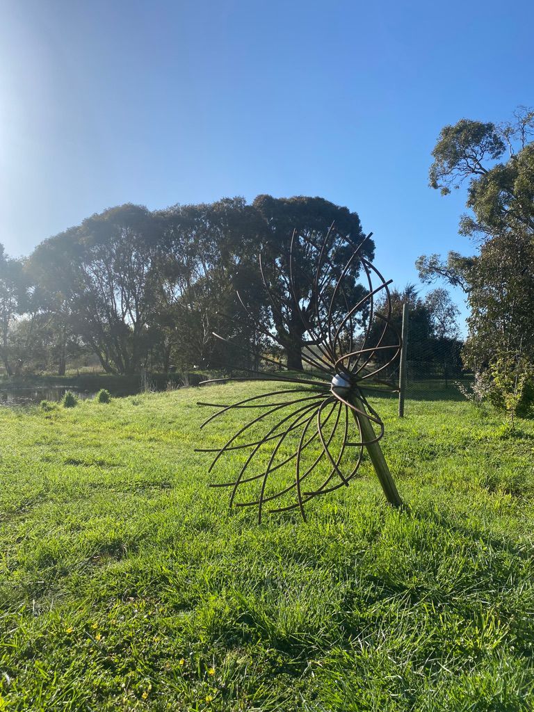 Metal sculpture of a stylized flower in a grassy field with trees under a blue sky.