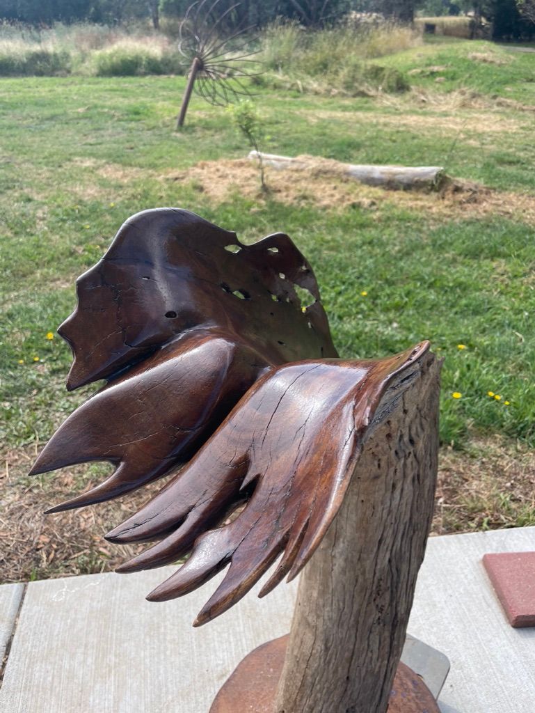Bronze abstract sculpture with wing-like elements on a wooden post, set in a grassy outdoor area.