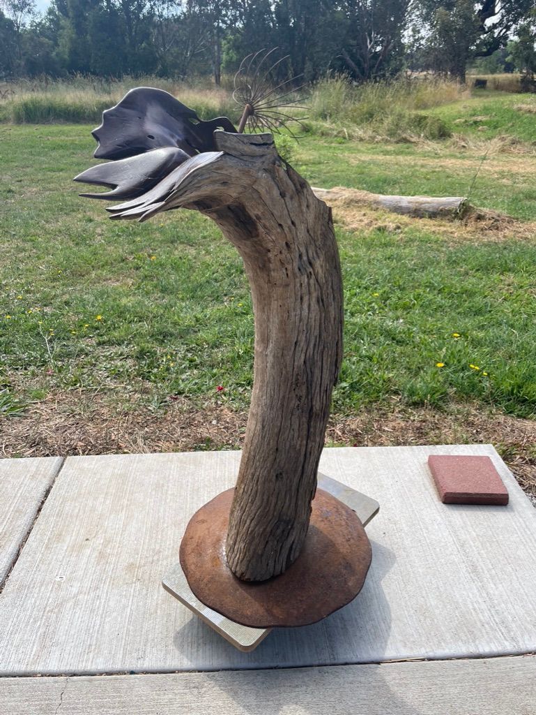 Sculpture of a weathered, curving tree trunk with abstract wing-like forms, set on a stone base, outdoors.