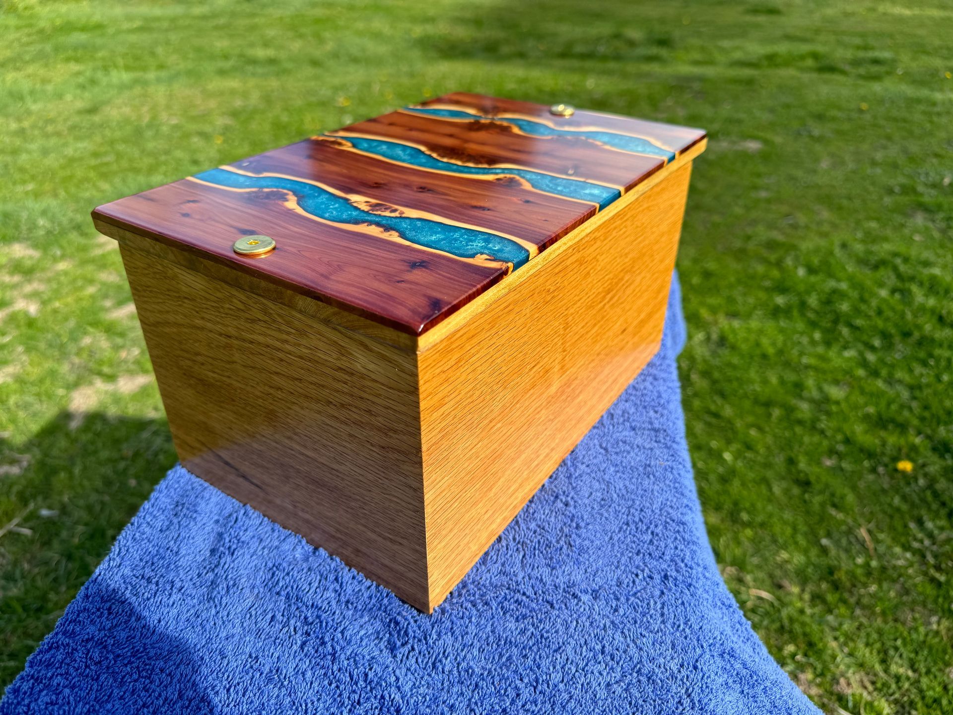 Wooden box with a river-like design of turquoise resin set into the lid, on a blue towel in a grassy setting.