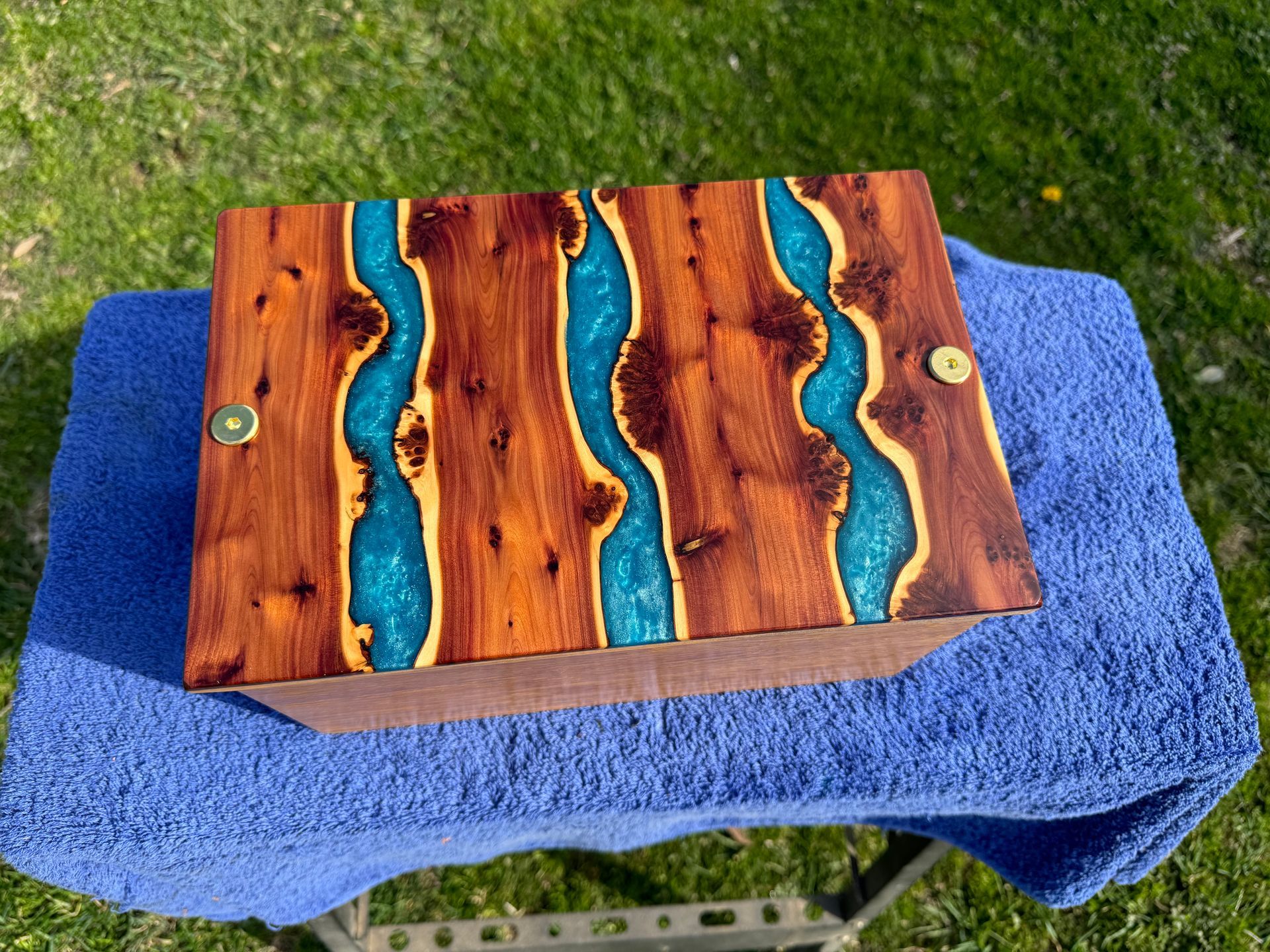 Wooden box with river-like blue epoxy resin design on the lid, resting on a blue towel outdoors.
