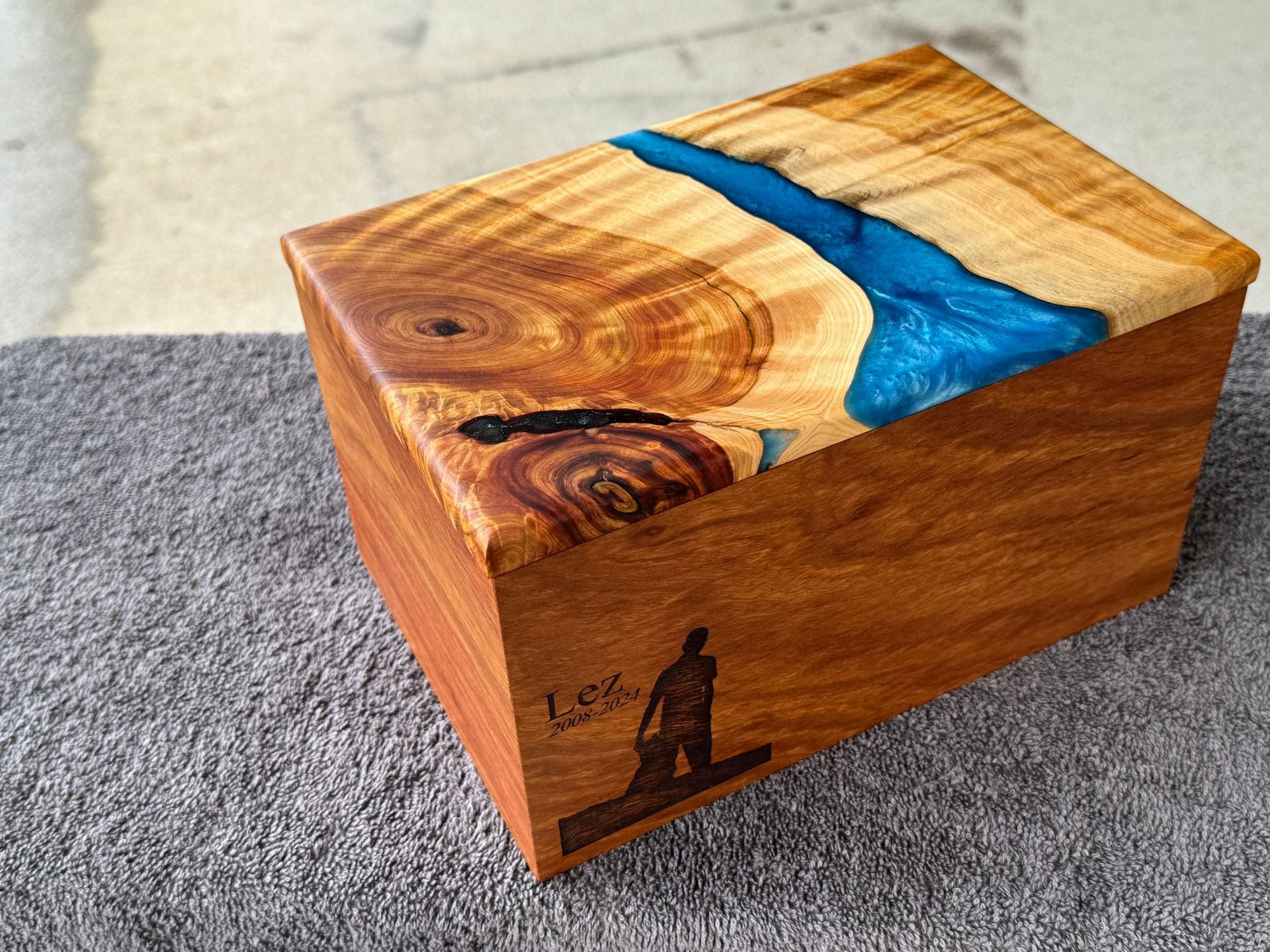 Wooden box with a blue resin river on the lid, etched surfer and signature on the side.