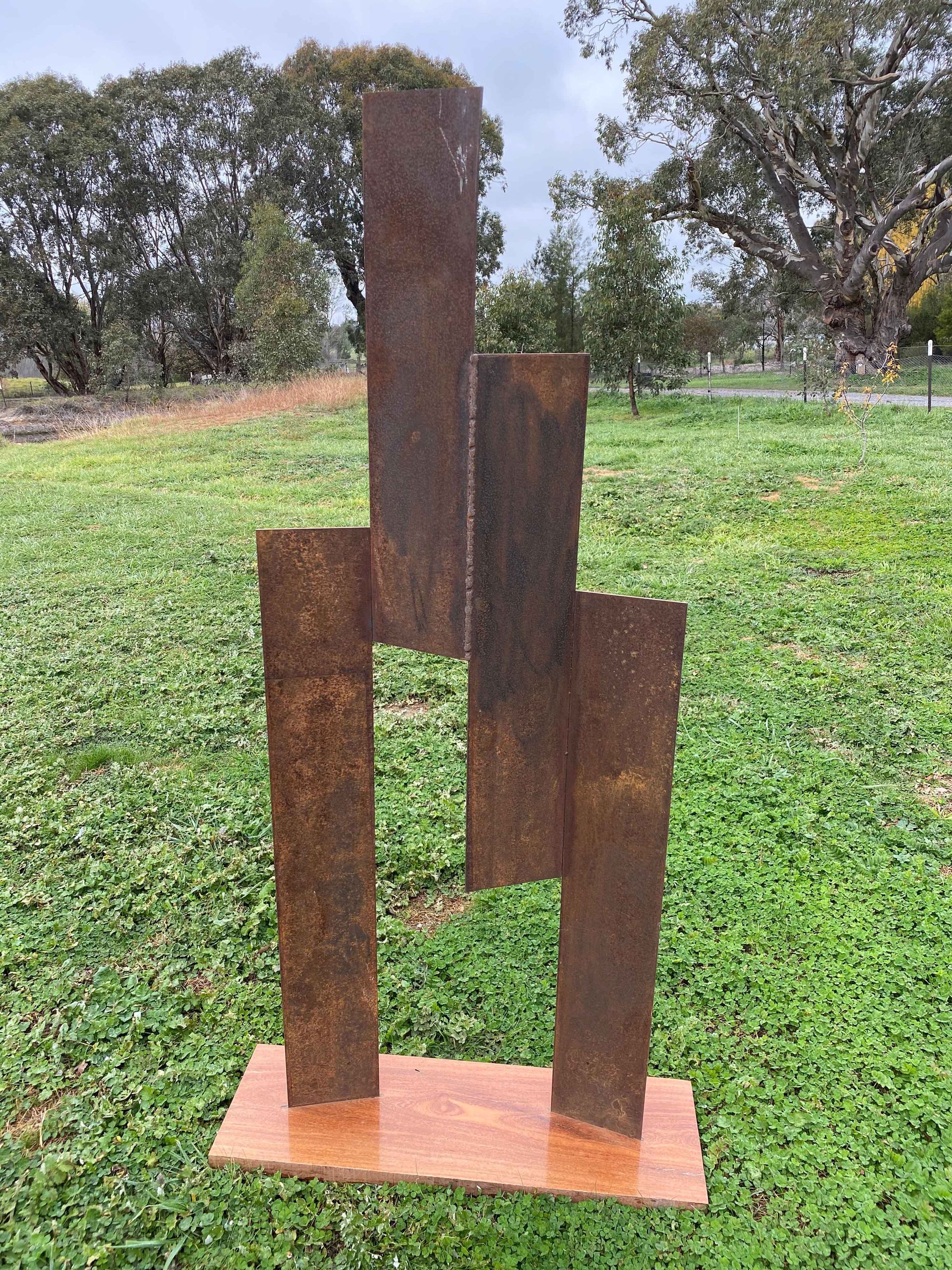 Rust-colored, abstract metal sculpture with rectangular uprights on a rectangular base; set in a grassy outdoor area.