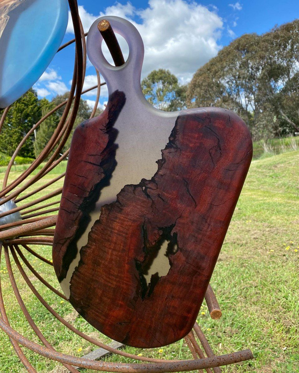 Wooden cutting board with reddish-brown wood and a gray resin river, handle on top, displayed outdoors.