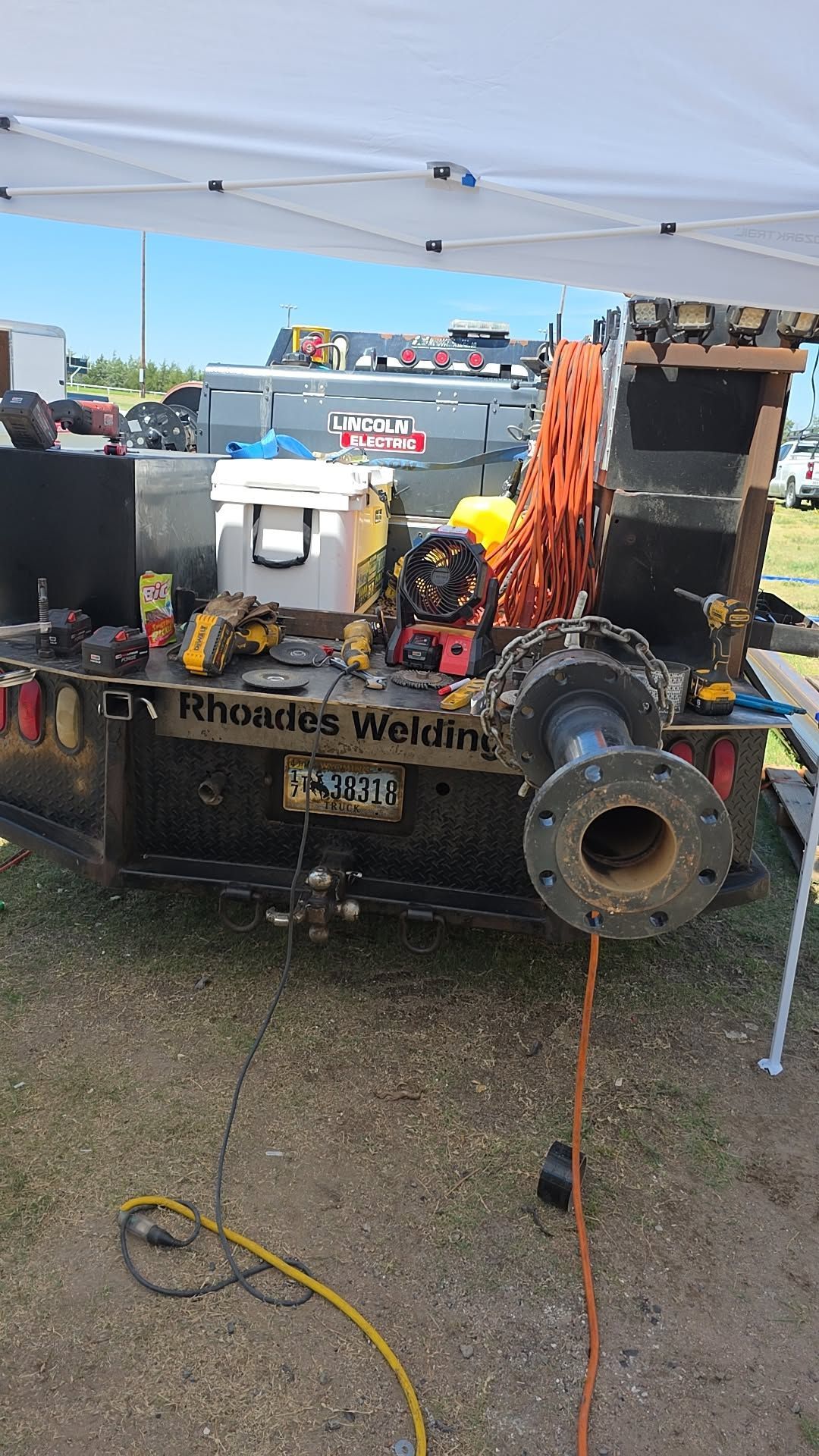 The back of a welding truck with tools, a cooler, and a metal pipe flange mounted on the bumper under a white canopy.
