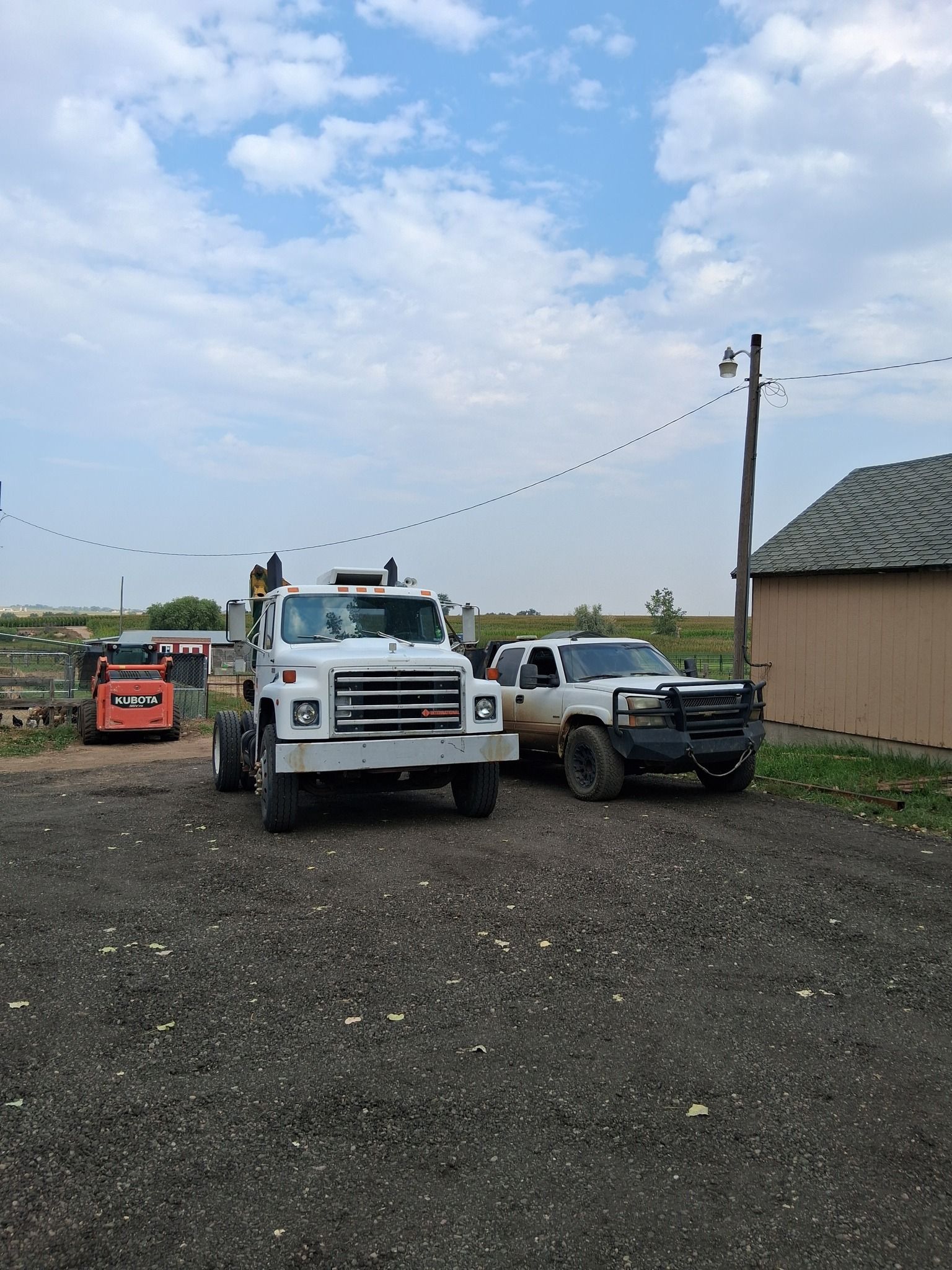 A white semi-truck, a silver pickup, and an orange skid steer parked on a gravel lot next to a small building.
