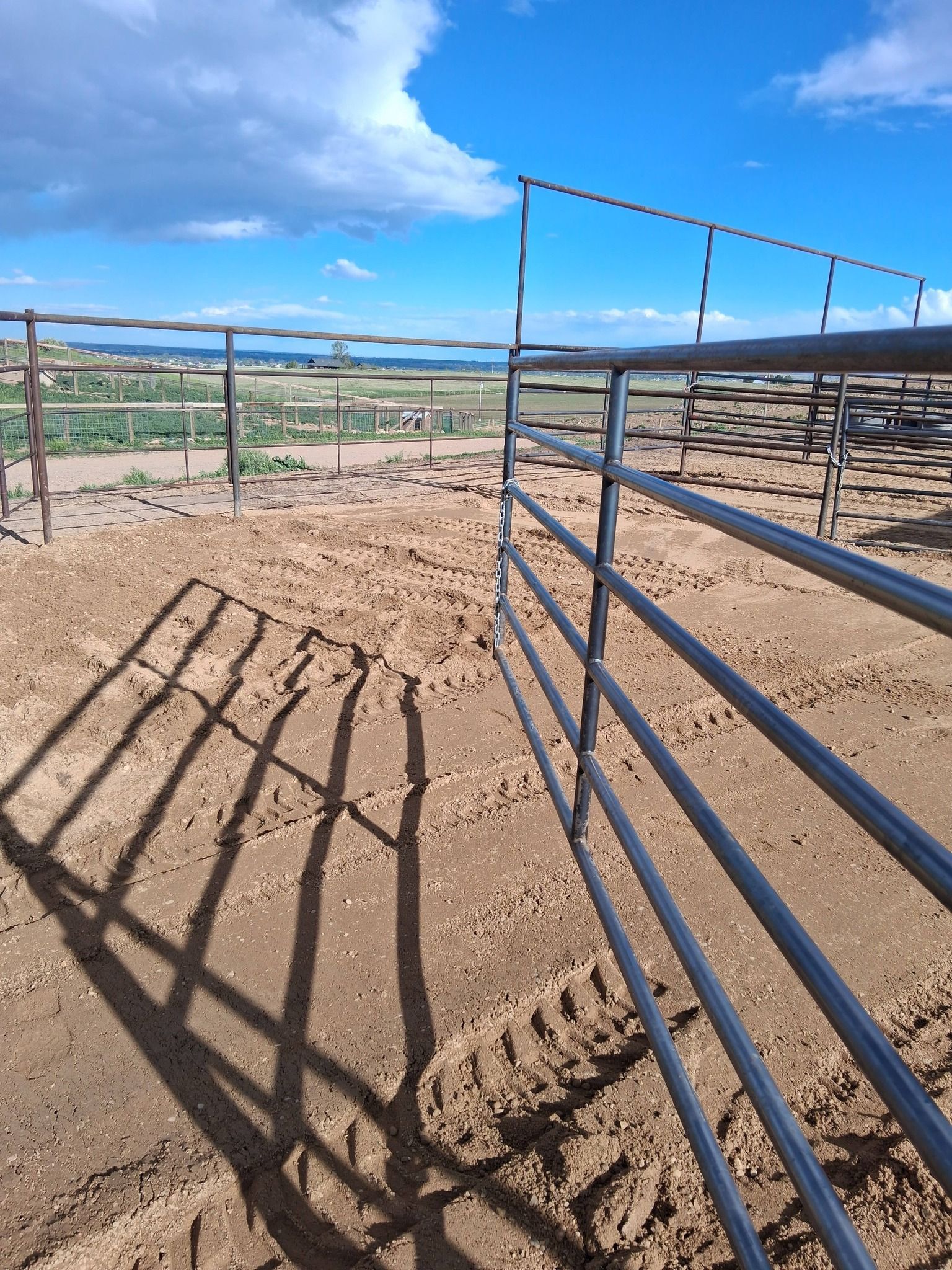 Metal livestock corral panels and their shadows on dirt ground under a blue, partly cloudy sky.