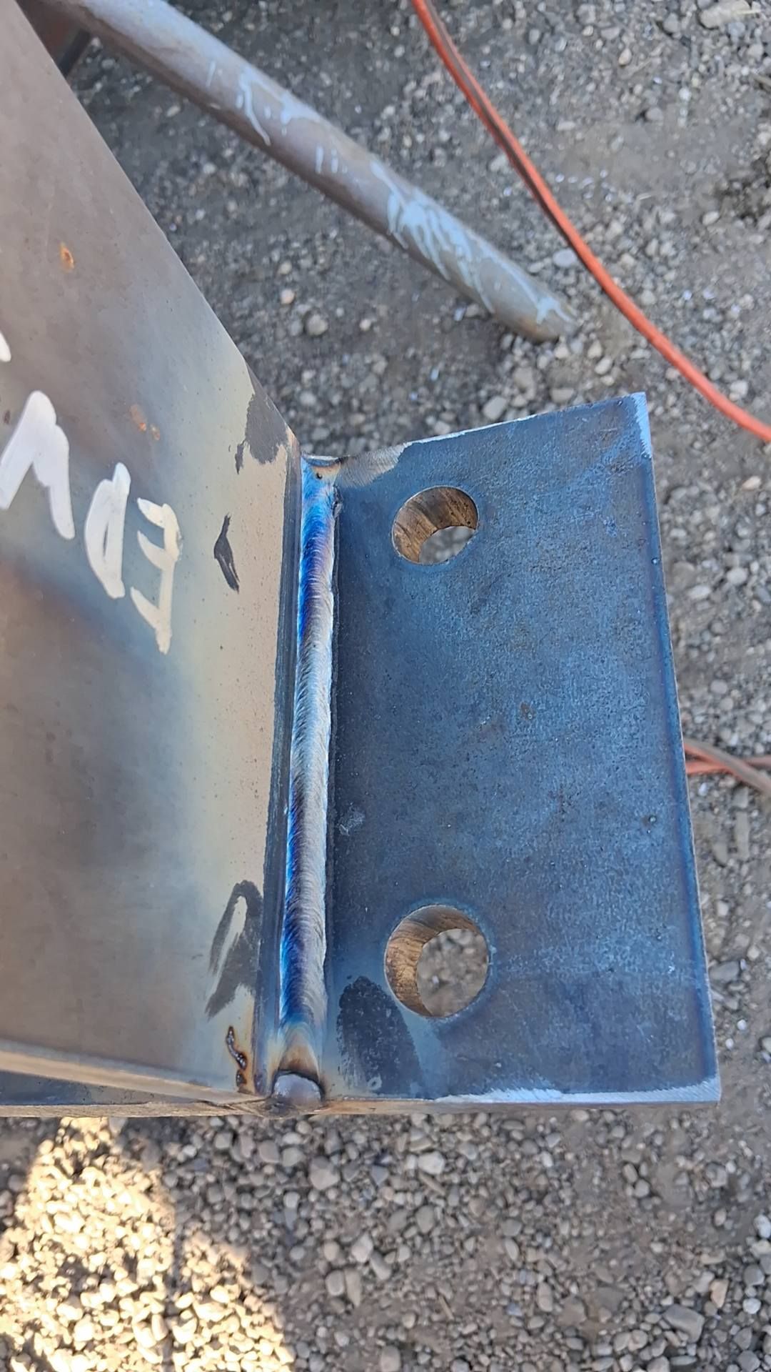 A steel bracket welded to a metal beam, featuring two circular bolt holes and a vertical weld seam.
