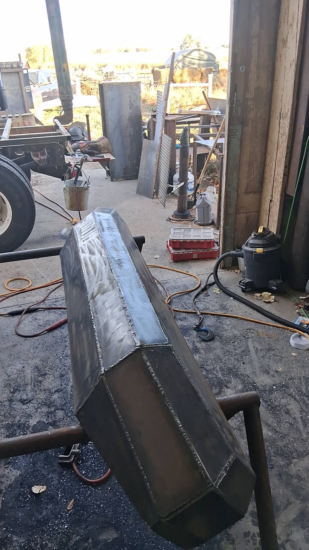 A welded, multi-faceted metal bumper rests on a stand in a workshop with tools and a vehicle frame in the background.