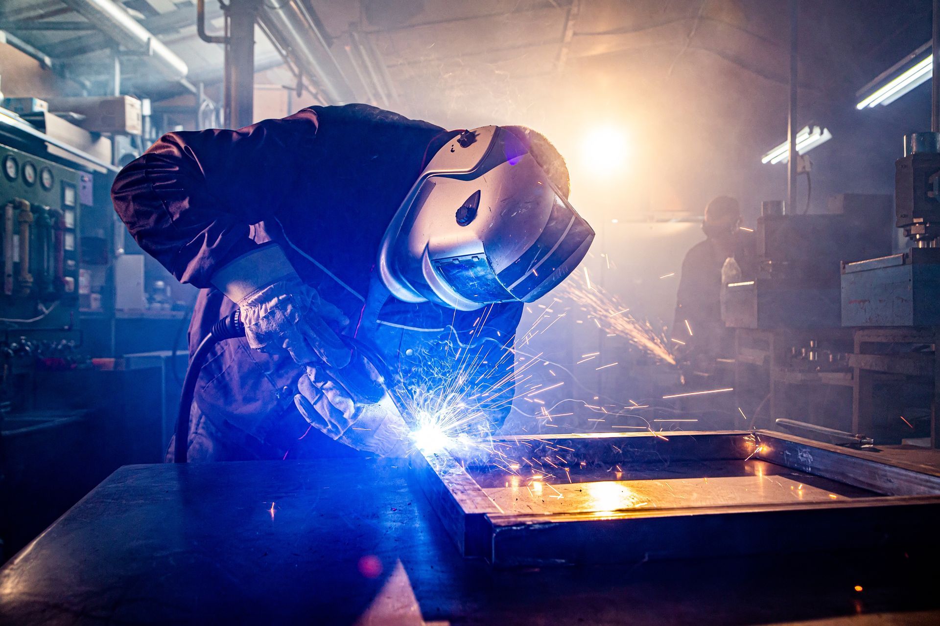 A welder in protective gear uses a torch to join metal, creating bright sparks and a blue glow in a dimly lit workshop.