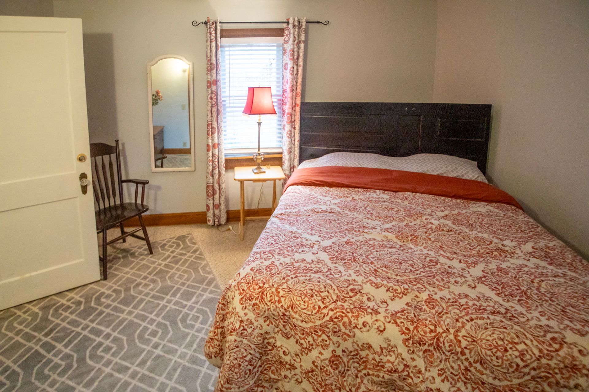 Bedroom with bed, small table with lamp, chair, mirror, window with curtains.