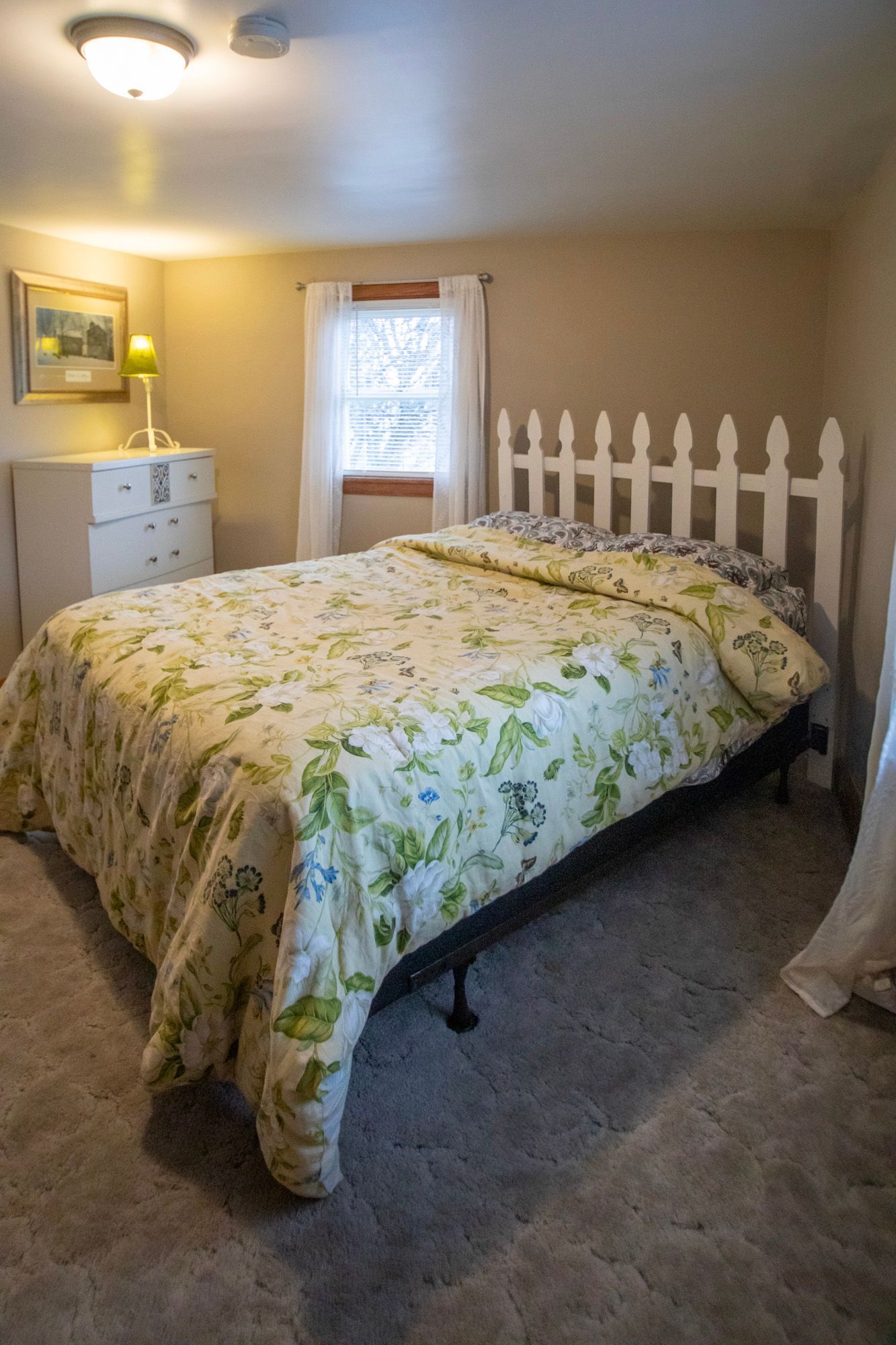 Bedroom with a bed, dresser, window, and picket fence headboard. Floral bedding and neutral walls.