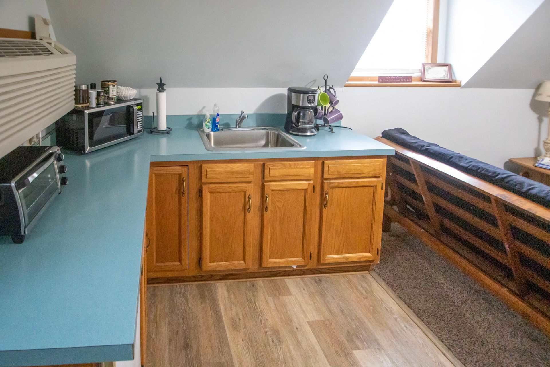 Small kitchen area with blue counter, sink, microwave, cabinets, and a window.