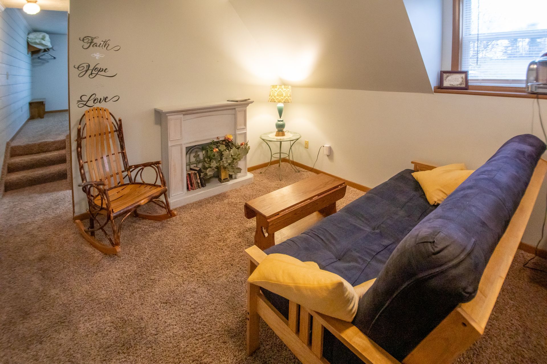 Cozy living room with futon, rocking chair, faux fireplace, and wall art. Brown carpet, soft lighting.