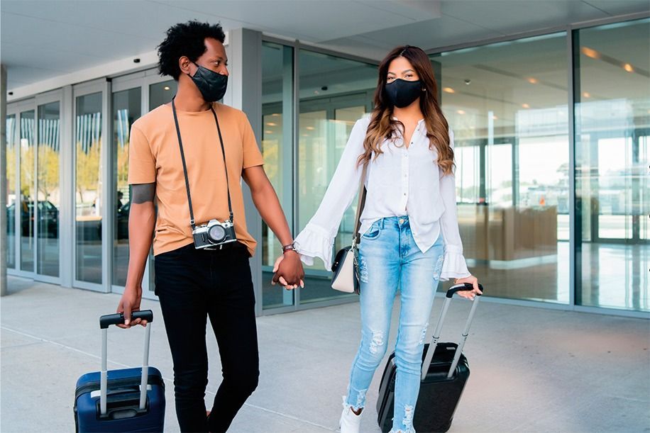 Casal caminhando de mãos dadas, puxando malas, usando máscaras perto de portas de vidro.