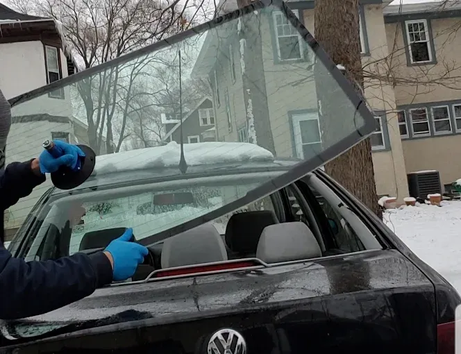 Person removing a car's rear window. Black car, blue gloves, suction cup, snowy background, holding window.