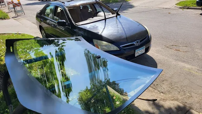 A black car with its windshield broken, new windshield in front, outdoors on a sunny day.