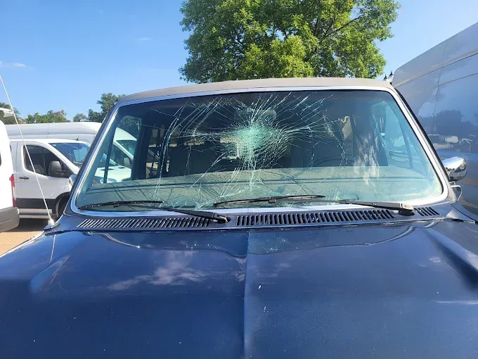 A dark blue car's windshield is shattered. The car is outdoors, parked between two white vans, under a blue sky.