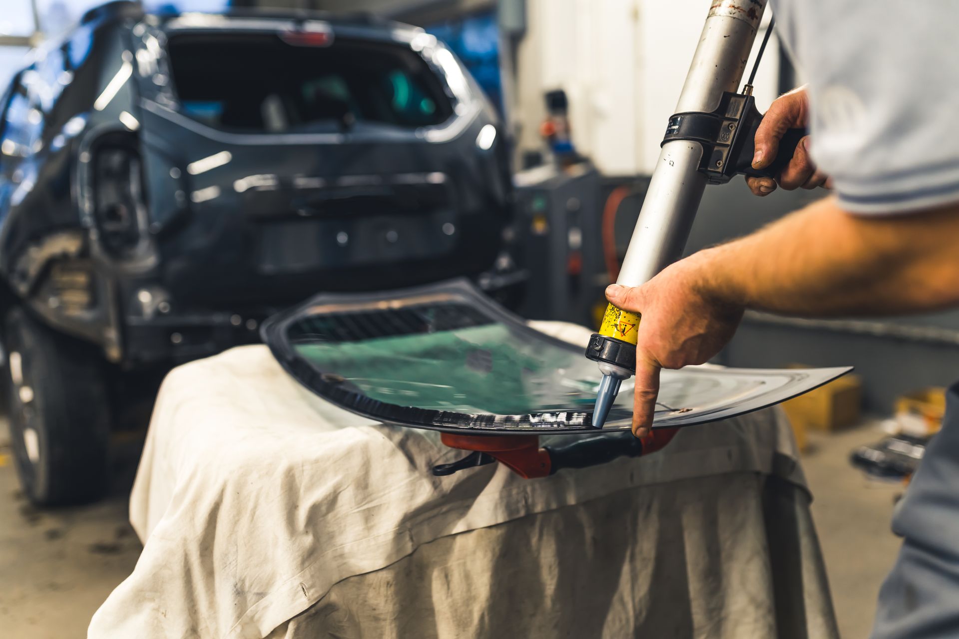 A mechanic applying sealant around a car windshield. The car is black and inside a repair shop.