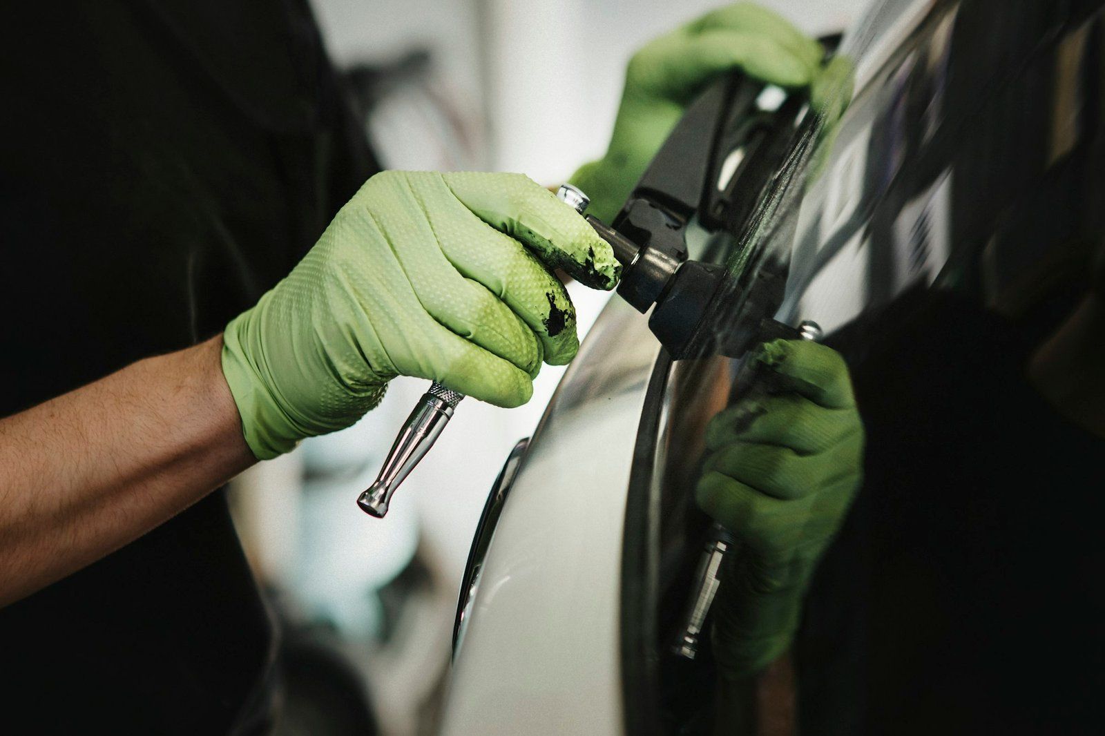 Hands wearing green gloves spraying paint on a car.
