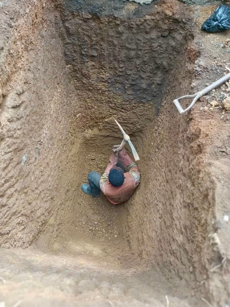 Person in a deep trench, using a pickaxe on the wall. Beige soil, shovel nearby.