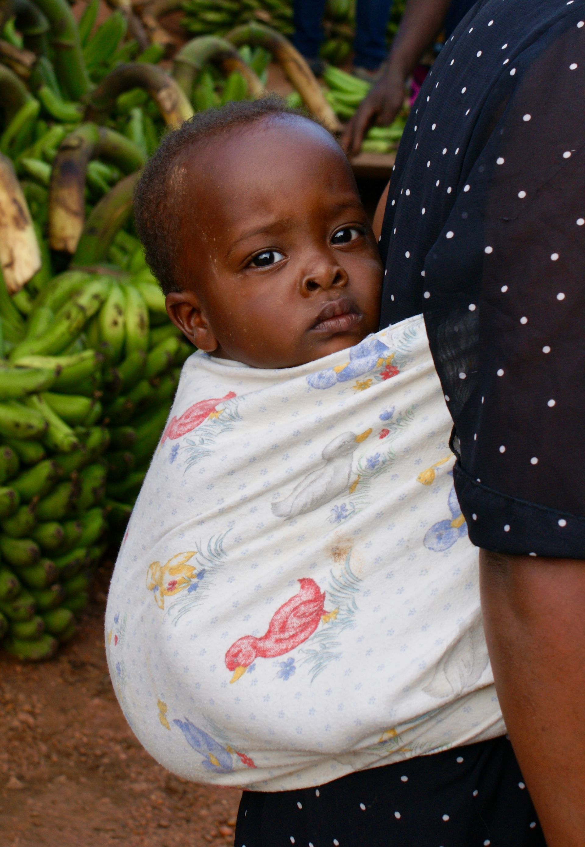 Baby in patterned wrap against a person's torso, bananas in background.