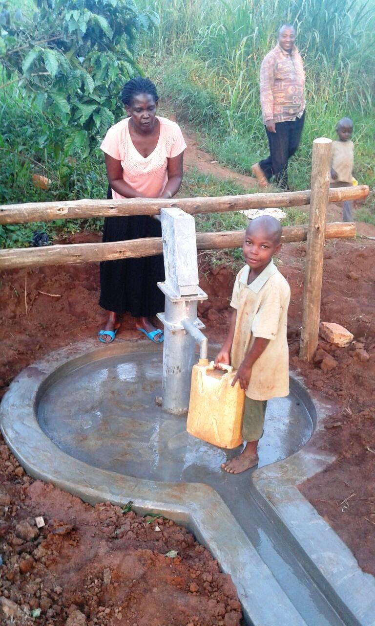 Child pumps water into a container at a well; two adults watch. Outdoors, dirt ground, concrete well surround.