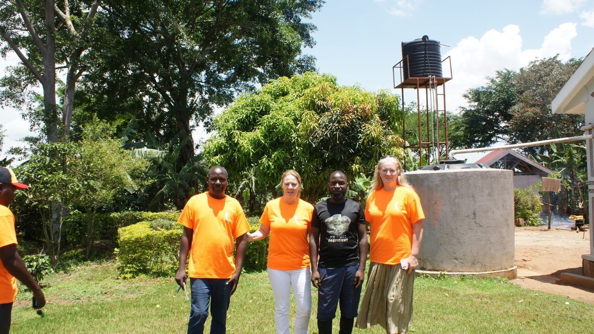 Four people wearing orange shirts stand outside near a water tank in a grassy area.