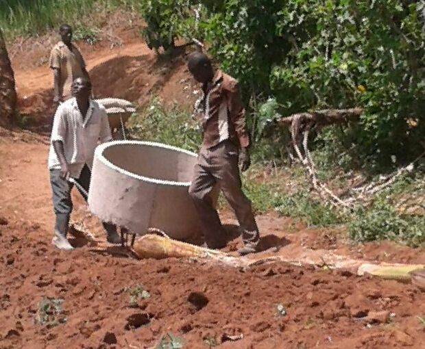 Men carrying a large concrete ring on a dirt path near trees.