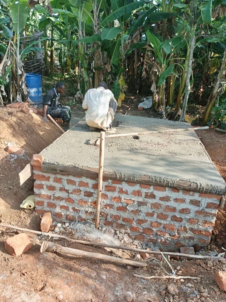 Construction workers building a rectangular brick structure with a concrete top, outdoors near banana trees.